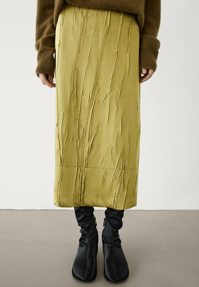 Personne portant une jupe midi vert olive froissée, un pull marron avec des poignets allongés, et des bottines noires décontractées, debout sur un sol gris.