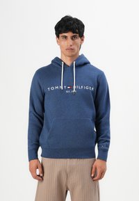 Man in een blauwe Tommy Hilfiger hoodie met een zak voorop en beige broek, staand tegen een effen witte achtergrond.