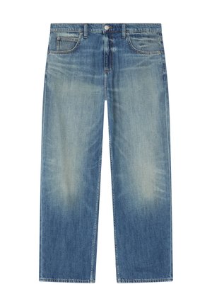 Junger Mann trägt ein weißes T-Shirt, ein blaues Jeanshemd, eine cremefarbene Sherpa-Weste, lockere blaue Jeans und weiße Sneakers und schaut zur Seite.