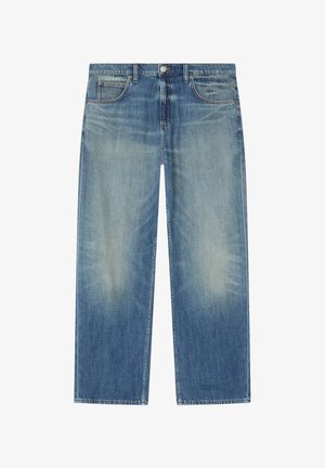 Junger Mann trägt ein weißes T-Shirt, ein blaues Jeanshemd, eine cremefarbene Sherpa-Weste, lockere blaue Jeans und weiße Sneakers und schaut zur Seite.