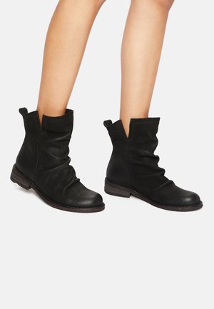 Jambes portant des bottines noires texturées à la cheville avec tige souple, talon bas empilé et languettes de traction sur fond blanc.