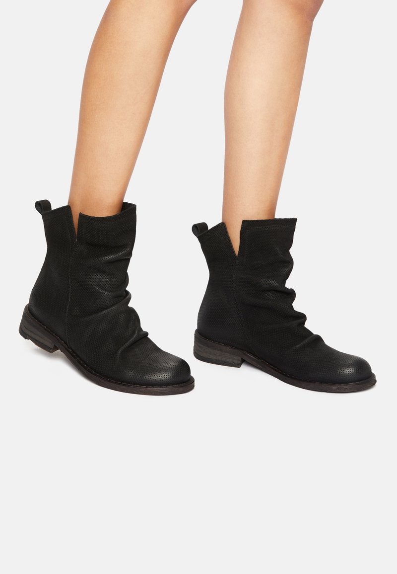 Jambes portant des bottines noires texturées à la cheville avec tige souple, talon bas empilé et languettes de traction sur fond blanc.