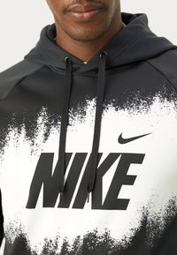 Svart hettegenser med hvitt sprut-design og stor svart "NIKE"-logo. Har snøring i halsen, myk tekstur og ribbestrikkede kanter.