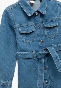 Vaaleansininen farkkumekko, jossa on kaulus, pitkät hihat, kaksi rintataskua, etupainatusnapit ja vyö vyötäröllä. Teksturoitu kangas.