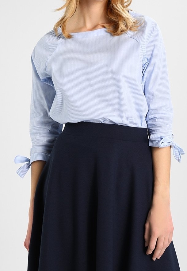 Blouse rayée bleu clair à manches trois-quarts, avec des accents en lien aux poignets. Associée à une jupe évasée navy foncé.
