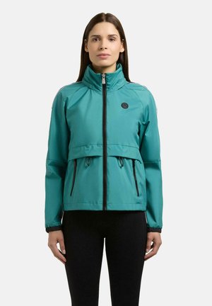 Kobieta w turkusowej bluzie zapinanej na zamek z wysokim kołnierzem oraz w czarnych spodniach, stojąca przodem na jednolitym tle.