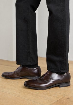 Braune Leder-Dressschuhe mit glatter Textur, spitzem Zeh und dunkelburgunderfarbenen Schnürsenkeln. Kombiniert mit schwarzen, schmal zulaufenden Hosen.