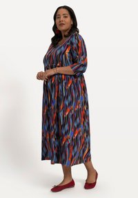 Maxikleid mit einem spiralförmigen Muster in Blau, Orange und Rot. Glatter Stoff mit einem runden Ausschnitt und Dreiviertelärmeln. Getragen mit roten Ballerinas.