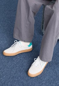 White sneakers with beige rubber soles and turquoise heel accents. Laces are white, and they are paired with loose gray pants on a blue carpet.