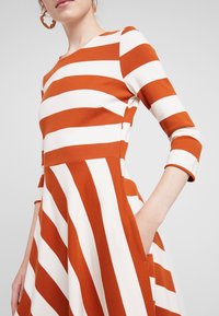 Robe rayée à manches longues, ornée de larges rayures orange et blanches, avec un corsage ajusté, une jupe évasée et des poches latérales. Tissu en mélange de coton.