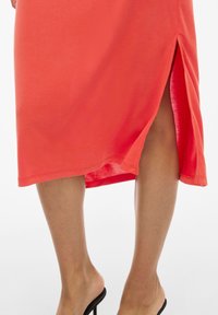 Vestido midi coral con una abertura lateral, presenta una textura suave y un dobladillo simple. Combinado con tacones abiertos negros.