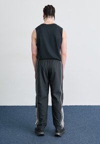 Cămașă fără mâneci de culoare neagră și pantaloni sport negri, cu dungi laterale albe. Persoana poartă încălțăminte întunecată, texturată. Fundal simplu.