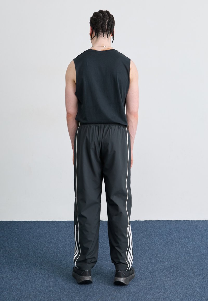 Cămașă fără mâneci de culoare neagră și pantaloni sport negri, cu dungi laterale albe. Persoana poartă încălțăminte întunecată, texturată. Fundal simplu.