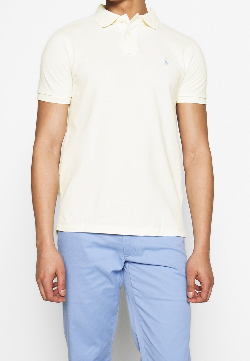 Muž v bledě žlutém polo tričku s krátkým rukávem a malou vyšitou nášivkou, oblečený v světlých modrých kalhotách, stojící na pozadí bílé zdi.