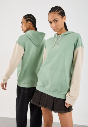 To personer med fletter som har på seg grønne hettegensere med beige ermer, en som vender foran i skjørt og den andre som vender bort i mørke bukser.