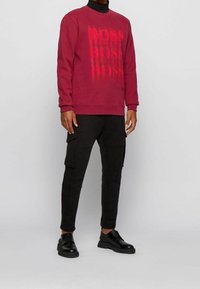 Felpa bordeaux con logo 'BOSS' nelle varie dimensioni sul davanti. Abbinata a pantaloni cargo neri e scarpe stringate nere con una leggera piattaforma.
