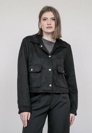 Jeune femme aux cheveux lisses mi-longs, portant une veste noire à boutons avec poches à rabat à l'avant et un pantalon noir, debout devant un fond neutre.