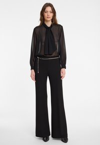 Transparante zwarte blouse met een strik, gecombineerd met zwarte hoge taille wijde broek. Accentuatie met een kettingriem voor extra detail. Soepele textuur, minimalistisch ontwerp.