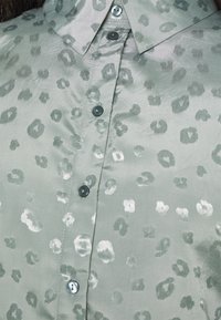 Lysegrøn skjorte med knapper, der har en skinnende, glat tekstur og leopardmønster i mørkere og lysere nuancer af grøn. Har rund krave og knapper.