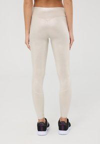 Leggings lucidi beige con vita alta, texture liscia e design aderente. Abbinati a scarpe da ginnastica nere con accenti viola.