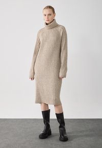 Beige bordázott kötött garbós ruha hosszú ujjal, térd alattig érő, fekete, térd fölé érő, vastag sarkú csizmával párosítva. Egyszerű, textúrázott dizájn.