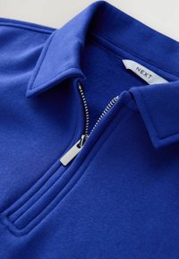 Kraag en halve rits op een blauwe fleece top met zichtbaar merklabel binnenin de kraag.