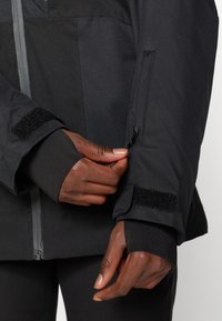 Casaco preto para exterior com tecido texturizado, velcro no pulso e bolso com zipper. Close-up de uma mão ajustando a manga.