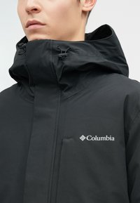Muškarac nosi crnu Columbia jaknu s visokim ovratnikom i podesivom kapuljačom, na kojoj se vidi brend logo na prsima.