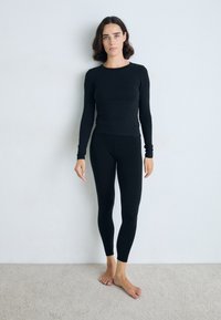 Zwarte langekorf top en leggings gemaakt van een zachte, rekbare stof. Strak ontwerp met naadloze randen. Model staat blootsvoets op het tapijt.