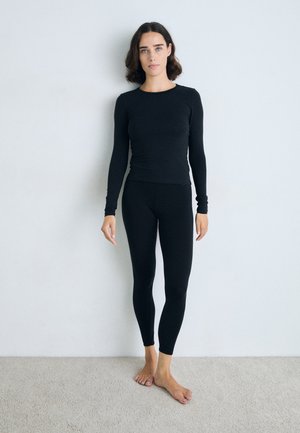 Top nero a maniche lunghe e leggings realizzati in un tessuto morbido e elasticizzato. Design aderente con bordi senza cuciture. Modella in piedi a piedi nudi su un tappeto.