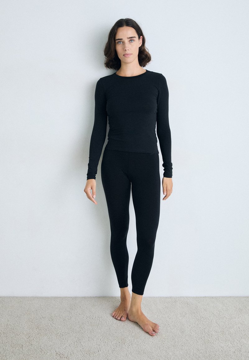 Zwarte langekorf top en leggings gemaakt van een zachte, rekbare stof. Strak ontwerp met naadloze randen. Model staat blootsvoets op het tapijt.
