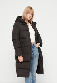 Cappotto imbottito nero con cappuccio, caratterizzato da un design a righe orizzontali, chiusura frontale con zip e due tasche laterali. Indossato sopra un maglione bianco.