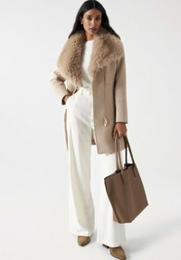 Manteau en laine beige avec un col en fourrure, fermeture éclair et poches latérales, associé à un pantalon blanc à jambes larges et un sac fourre-tout marron.