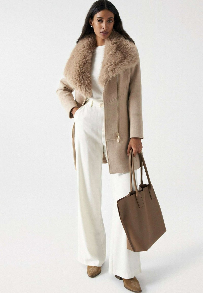 Manteau en laine beige avec un col en fourrure, fermeture éclair et poches latérales, associé à un pantalon blanc à jambes larges et un sac fourre-tout marron.