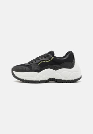 Czarno-biała masywna sneaker z grubą, teksturowaną podeszwą, czarnymi sznurowadłami, panelami z siatki i żółtym brandingiem Armani Exchange z boku.