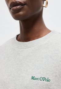 Κοντινό πλάνο ενός ατόμου που φοράει ανοιχτό γκρι φούτερ Marc O'Polo και χρυσό σκουλαρίκι, εμφανίζοντας τα χείλη και τον λαιμό.