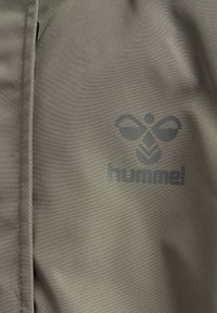 Sportowa marynarka z tkaniny khaki o gładkiej fakturze, z ciemnoszarym tłoczonym logo i przeszyciami wzdłuż krawędzi.