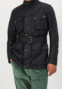 Laivastonsininen vedenpitävä takki, jossa on korkea kaulus, neljä etutaskua, vyö vyötäröllä ja metalliset napit koristelussa. Sileä pinta.