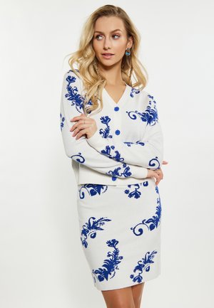 Frau mit langen blonden Haaren, die ein weißes zweiteiliges Outfit mit blauen Blumenmustern trägt, steht mit verschränkten Armen und schaut zur Seite.