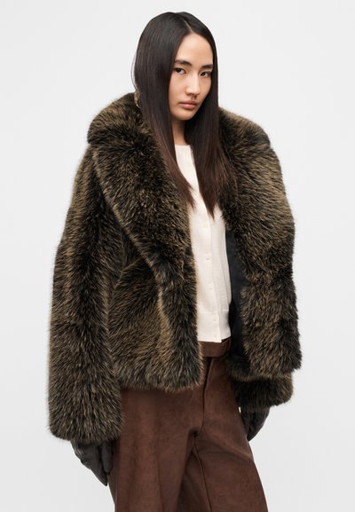 Καφέ μπουφάν από συνθετική γούνα με φαρδιά γιακά και μακριά μανίκια, σετ με ανοιχτό κρεμ τοπ και καφέ παντελόνι. Λιτή υφή, με όγκο σχεδίασης.