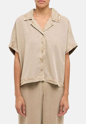 Frau trägt ein lockeres, kurzärmeliges beiges Hemd mit Kragen und passenden beigen hoch taillierten Hosen, stehend vor einem weißen Hintergrund.