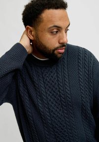 Navy knitted sweater with a textured cable pattern and ribbed cuffs. Features a high, rounded neckline. Fabric appears thick and cozy.