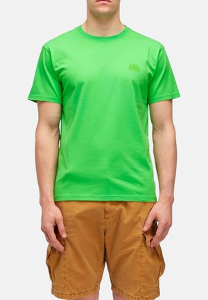 Uomo che indossa una maglietta verde brillante a maniche corte e pantaloncini cargo marroni, in piedi su uno sfondo chiaro e uniforme.