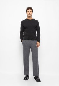 Szürke gyapjú keverék nadrág és fekete hosszú ujjú pulóver, fényes fekete cipővel viselve, fehér háttér előtt állva.