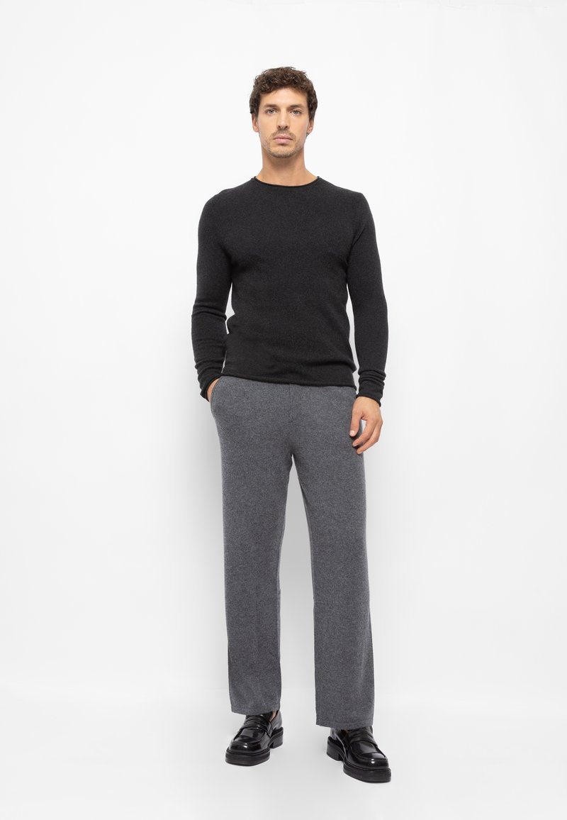 Szürke gyapjú keverék nadrág és fekete hosszú ujjú pulóver, fényes fekete cipővel viselve, fehér háttér előtt állva.