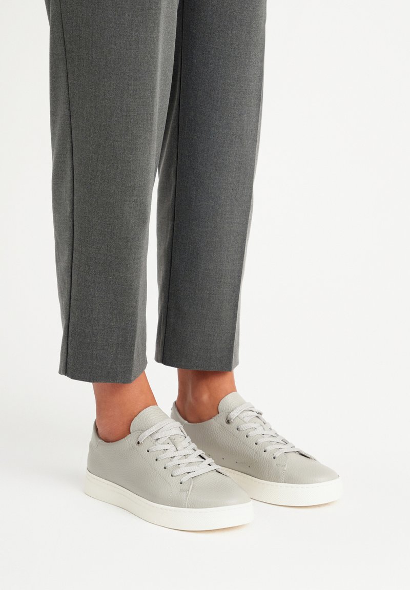 Sneakers in pelle grigia chiara con superficie testurizzata e punta tondeggiante, dotati di lacci piatti e suola in gomma bianca. Abbinati a pantaloni grigi su misura.