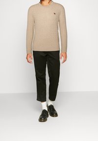 Muž oblečený v béžovém svetr s copánkovým vzorem, černé zkrácené kalhoty, bílé ponožky a černé šněrovací boty stojící před jednobarevným pozadím.