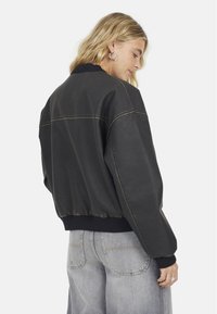 Giacca bomber nera con orlo e polsini a coste, caratterizzata da cuciture gialle in evidenza e una vestibilità rilassata. Indossata su jeans grigi chiari.