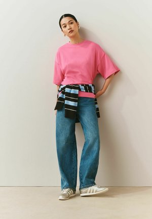 Femme portant un t-shirt oversize rose, un jean bleu, un pull rayé noué à la taille et des baskets blanches, debout contre un mur uni.