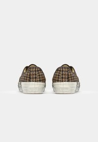 Brązowe i czarne buty typu slip-on w kratkę z teksturowaną cholewką, białą gumową podeszwą i zaokrąglonym noskiem.